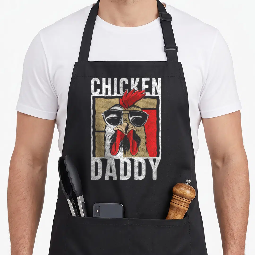Daddy rooster apron - Cartoon rooster head with sunglasses and CHICKEN DADDY text in white red tan grey — shown on a black cotton canvas apron against a white background