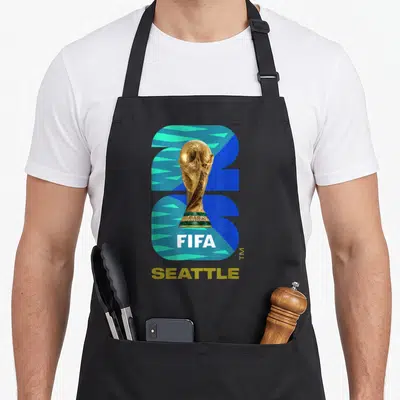 Fifa seattle apron - Gold trophy over blue teal background with white FIFA and gold Seattle text — shown on a black cotton canvas apron against a white background