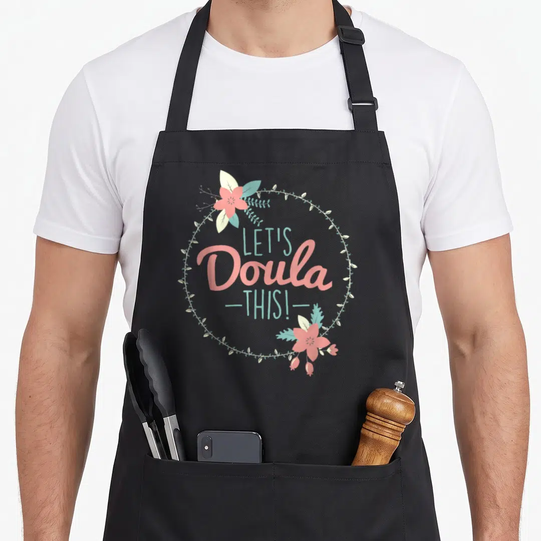 Midwife doula apron - LET'S Doula THIS! text inside a circular floral wreath with pink flowers and teal leaves — shown on a black cotton canvas apron against a white background