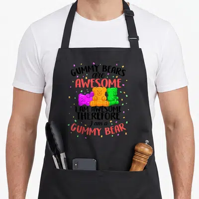 Dancing bear apron - Three orange, purple, and green gummy bears surrounded by confetti below stacked bold text reading — shown on a black cotton canvas apron against a white background