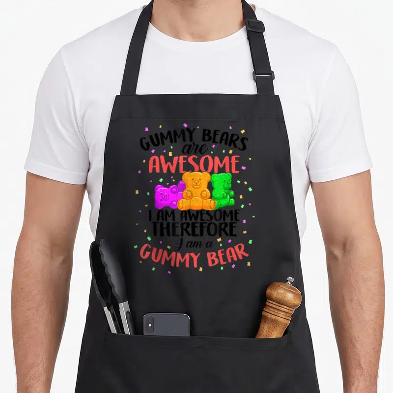 Dancing bear apron - Three orange, purple, and green gummy bears surrounded by confetti below stacked bold text reading — shown on a black cotton canvas apron against a white background