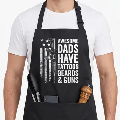Gun tattoo aprons - White rifle overlapping distressed flag next to stacked text reading AWESOME DADS HAVE TATTOOS — shown on a black cotton canvas apron against a white background