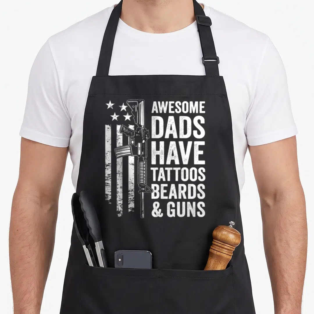 Gun tattoo aprons - White rifle overlapping distressed flag next to stacked text reading AWESOME DADS HAVE TATTOOS — shown on a black cotton canvas apron against a white background