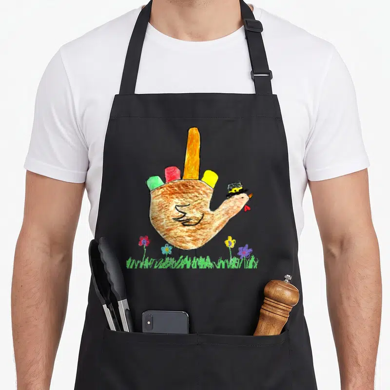 Friendsgiving pilgrim apron - Cartoon turkey in top hat with red, yellow, green, and red tail feathers standing on grass — shown on a black cotton canvas apron against a white background