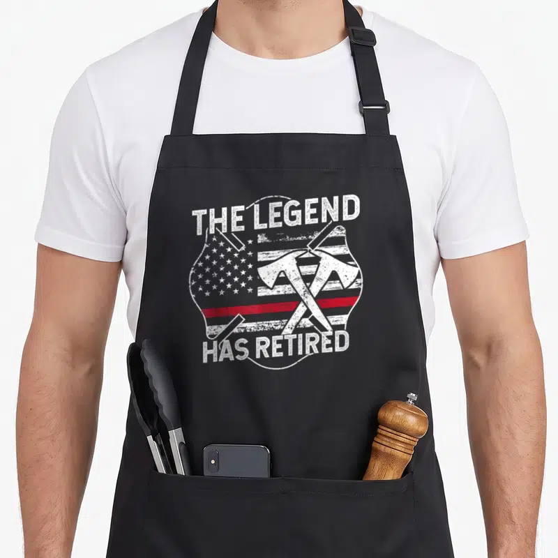 Legend retired firefighter cooking apron - Distressed text THE LEGEND HAS RETIRED over crossed axes and stylized flag — shown on a black cotton canvas apron against a white background