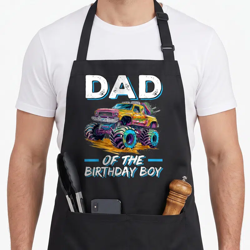 Monster bday truck apron - Cartoon monster truck in yellow blue pink and white with DAD OF THE BIRTHDAY BOY text — shown on a black cotton canvas apron against a white background