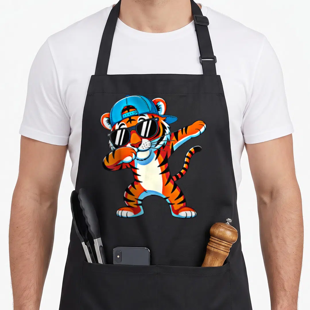 Dabbing animal print apron - Cartoon tiger dabbing in orange blue black with blue cap and sunglasses — shown on a black cotton canvas apron against a white background