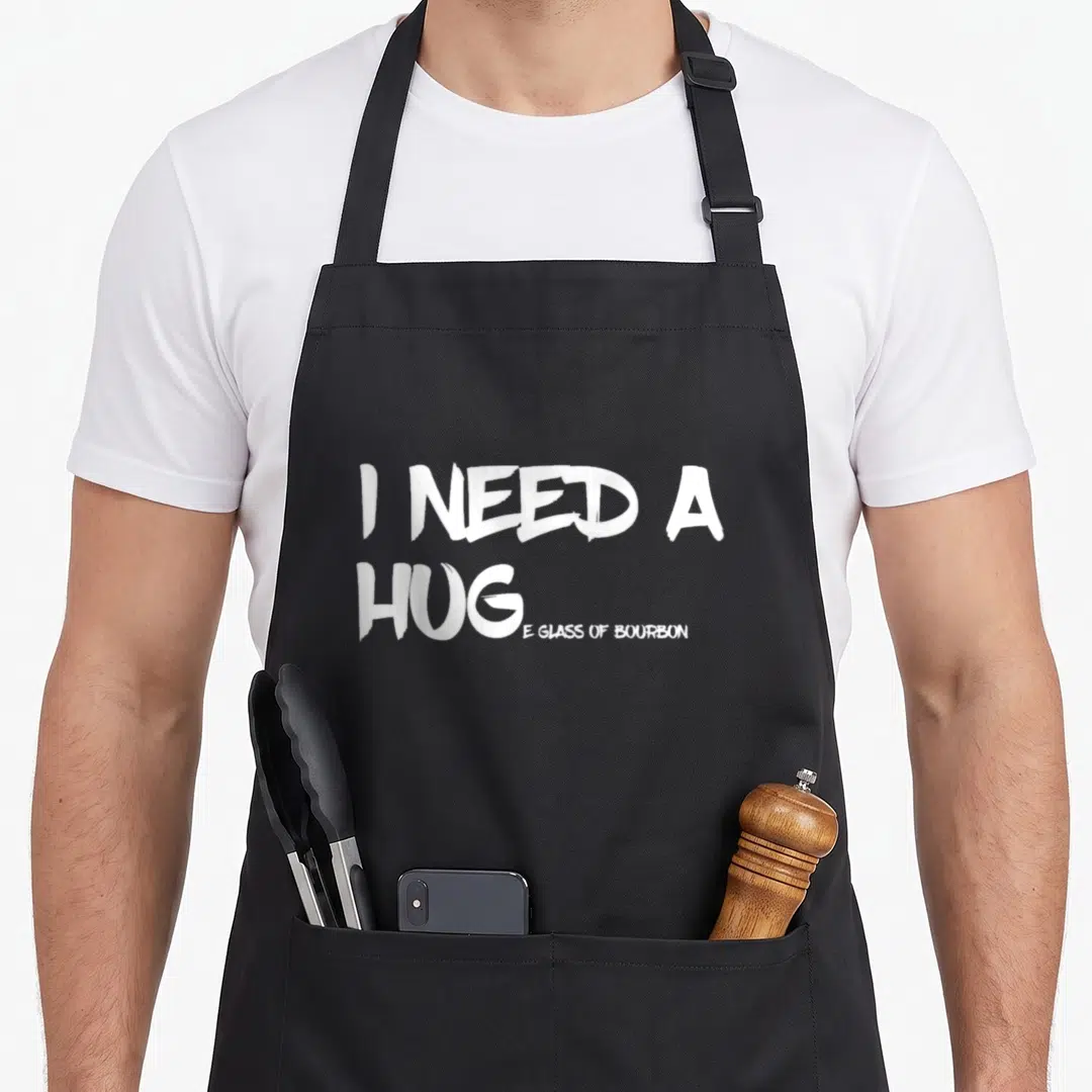 Drinking bourbon apron - White stacked typography reading I Need A Hug E Glass Of Bourbon on black fabric — shown on a black cotton canvas apron against a white background