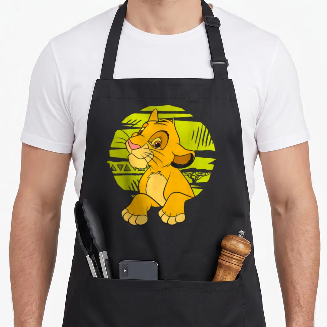 Simba 90s apron - Orange cartoon lion cub sitting in front of a yellow textured circle with tree silhouette — shown on a black cotton canvas apron against a white background