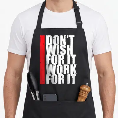 Motivation work hard apron - Stacked white text reading DON'T WISH FOR IT WORK FOR IT beside a vertical red bar — shown on a black cotton canvas apron against a white background