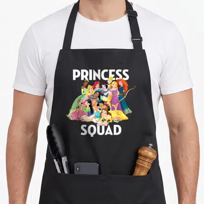 Squad princess apron - PRINCESS SQUAD text with clustered cartoon princesses in pink yellow blue green white — shown on a black cotton canvas apron against a white background