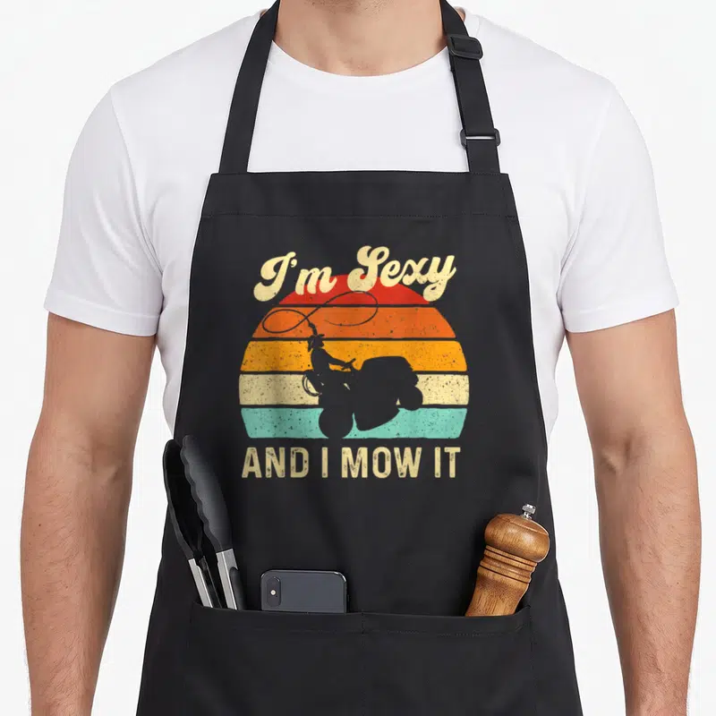 Mow trimmer apron - Retro cowboy on riding mower silhouette with I'm Sexy AND I MOW IT text over striped sun — shown on a black cotton canvas apron against a white background