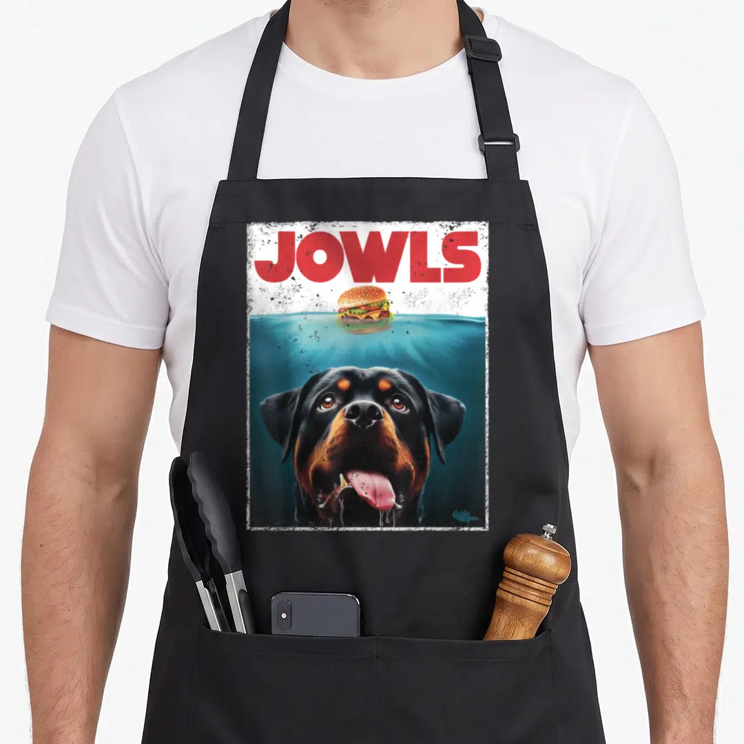 Burger dog apron - Red JOWLS text above a floating burger and submerged black and tan dog head — shown on a black cotton canvas apron against a white background