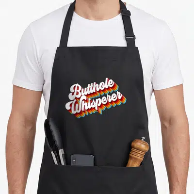 Butthole joke apron - White cursive text reading Butthole Whisperer with red orange yellow and blue 3D shadow layers — shown on a black cotton canvas apron against a white background