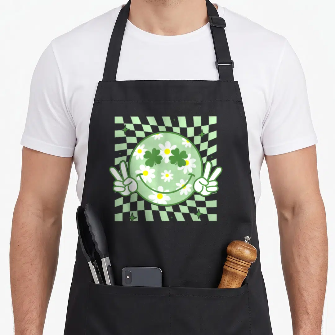 Patrick face on apron - Green smiley face with daisies and clovers flanked by two white peace sign hands over a wavy — shown on a black cotton canvas apron against a white background