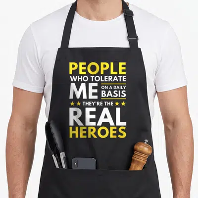 Tolerate daily apron - Stacked yellow and white text People Who Tolerate Me Real Heroes with four small stars — shown on a black cotton canvas apron against a white background