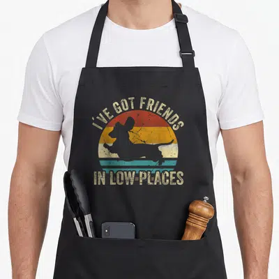 Friend dachshund wiener apron - Retro striped sun with dachshund silhouette and text I've Got Friends In Low Places — shown on a black cotton canvas apron against a white background