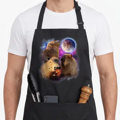 Stuffed animal apron - Three realistic capybaras under a moon with purple blue grey nebula — shown on a black cotton canvas apron against a white background