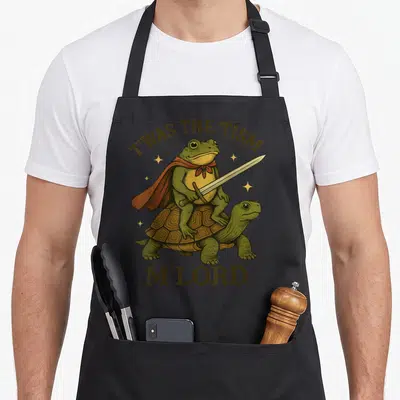 Twas frog aprons - Cartoon frog in red cape holding sword atop turtle with text T'WAS THE 'TISM M'LORD — shown on a black cotton canvas apron against a white background