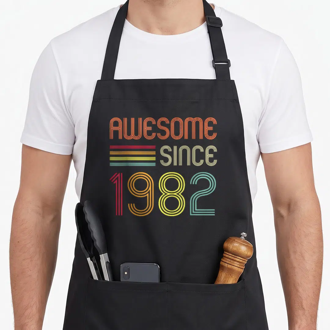42nd birthday apron - Retro AWESOME SINCE 1982 text with five colored horizontal stripes in orange, yellow, red, blue — shown on a black cotton canvas apron against a white background