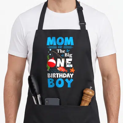 Ally birthday fish apron - MOM OF The Big ONE BIRTHDAY BOY text with fishing rod, red fish, orange starfish, and red — shown on a black cotton canvas apron against a white background