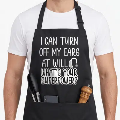Ear i turn grills on apron - Stacked white and black text reading I CAN TURN OFF MY EARS AT WILL with hearing aid icon — shown on a black cotton canvas apron against a white background