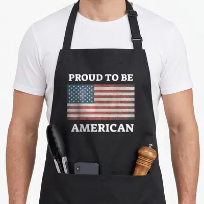 Patriotic proud american flag apron - Distressed USA flag centered between PROUD TO BE and AMERICAN text on black apron — shown on a black cotton canvas apron against a white background