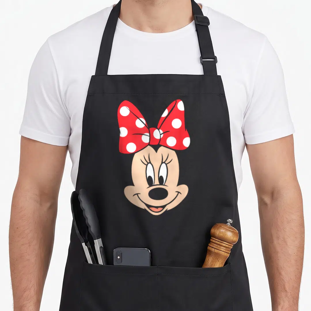 Dot bow apron - Minnie Mouse head with red white polka dot bow peach skin black outline cartoon — shown on a black cotton canvas apron against a white background