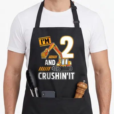 Vehicle 2nd apron - Cartoon excavator holding I'M sign next to number 2 with orange stripes and white text — shown on a black cotton canvas apron against a white background