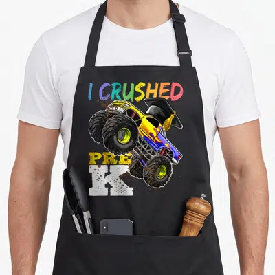 Pre truck apron - I Crushed Pre K monster truck with graduation cap and mud splatters — shown on a black cotton canvas apron against a white background