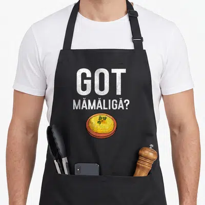 Mamaliga romanian apron - GOT MĂMĂLIGĂ? text above yellow bowl with green parsley on orange plate — shown on a black cotton canvas apron against a white background