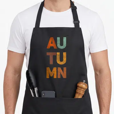 Timeless fall aprons - Stacked vintage typography reading AUTUMN in orange, sage, and rust block letters — shown on a black cotton canvas apron against a white background