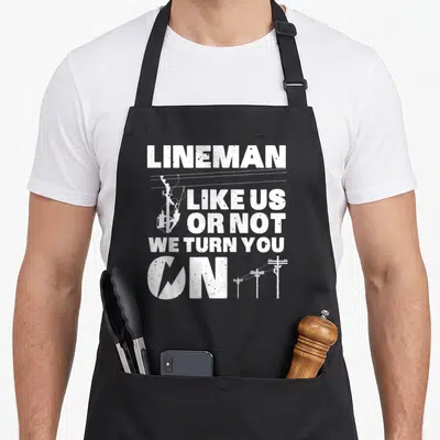 Lineman electronic electricians apron - White distressed text reading Lineman Like Us Or Not We Turn You — shown on a black cotton canvas apron against a white background