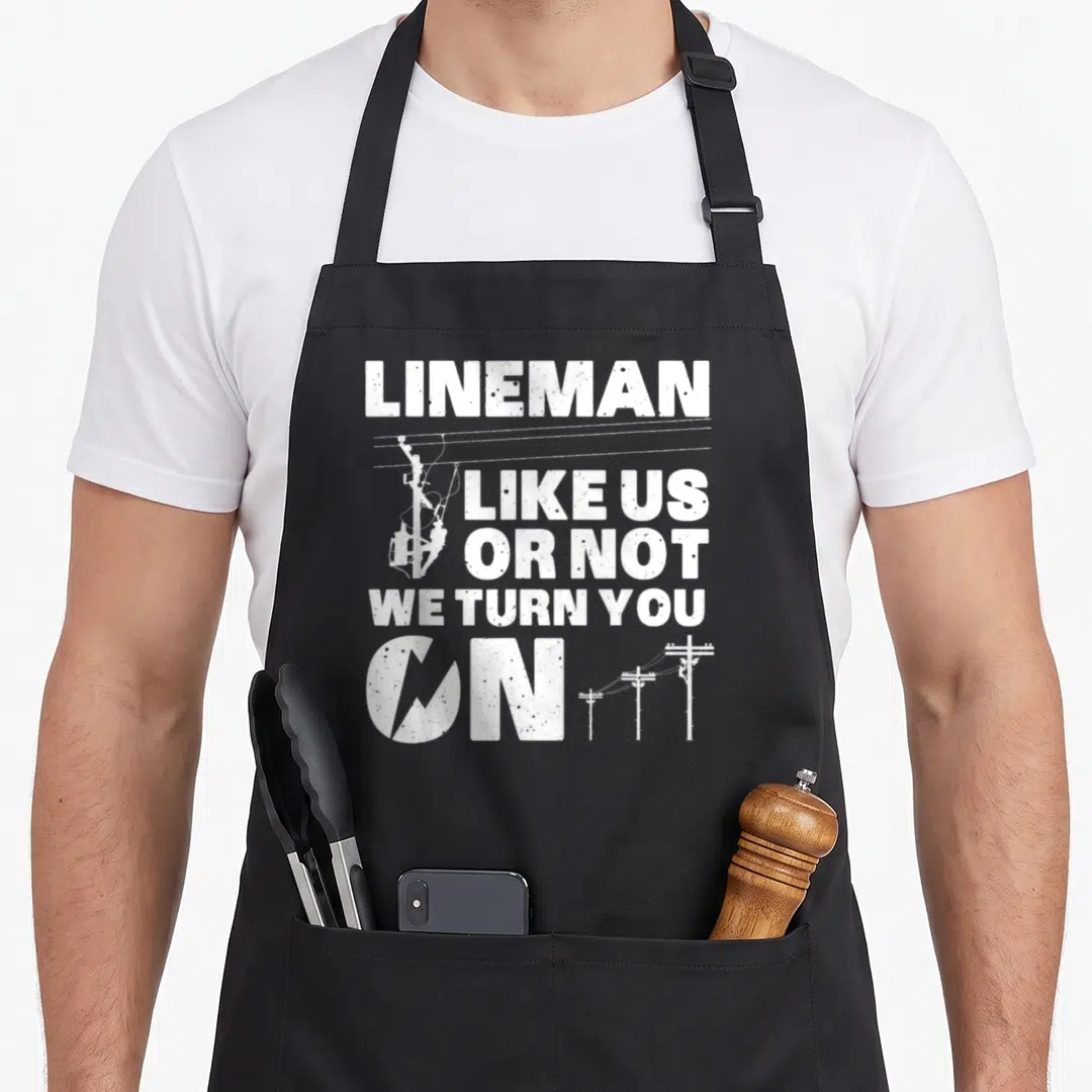 Lineman electronic electricians apron - White distressed text reading Lineman Like Us Or Not We Turn You — shown on a black cotton canvas apron against a white background