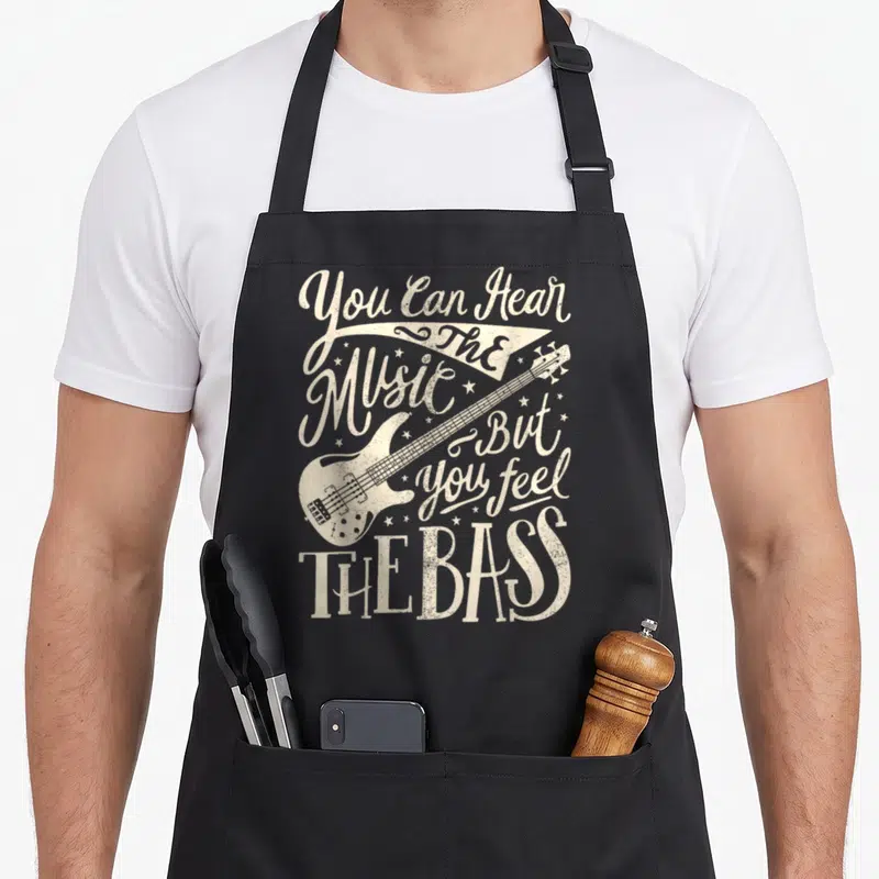 But bass apron - Beige bass guitar illustration with You Can Feel The Bass text and scattered stars — shown on a black cotton canvas apron against a white background