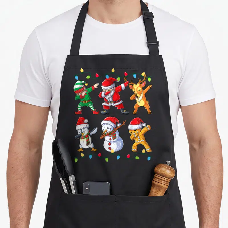 Dabbing christmas elf apron - Six cartoon Christmas characters dabbing under string lights in red green and brown — shown on a black cotton canvas apron against a white background