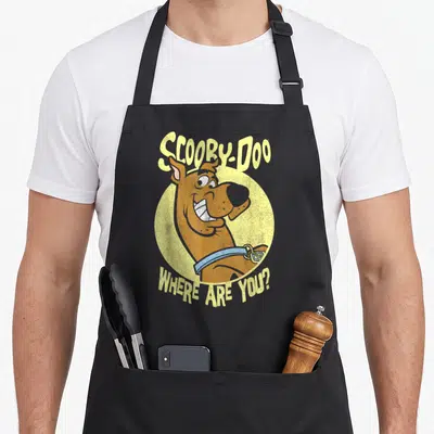 Cute decade apron - Cartoon brown dog in yellow circle with arched Scooby-Doo text — shown on a black cotton canvas apron against a white background