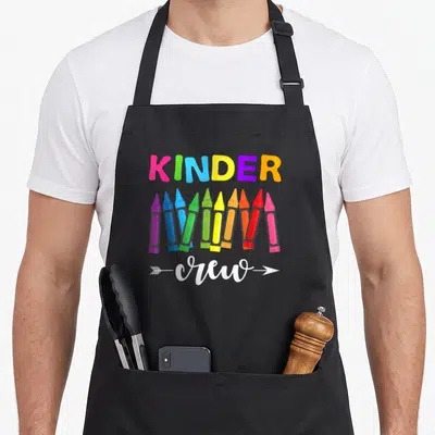 Kinder kindergarten teacher apron - KINDER crew text with eight colorful crayons and white arrowheads — shown on a black cotton canvas apron against a white background