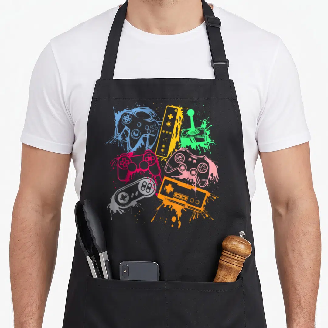 Console video game apron - Seven retro controllers and joystick in blue pink green orange purple over paint splatters — shown on a black cotton canvas apron against a white background