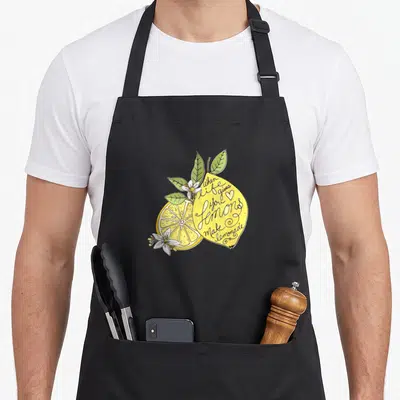 Give lemon apron - Cartoon lemon with black script text, green leaves, and white flowers on a yellow background — shown on a black cotton canvas apron against a white background