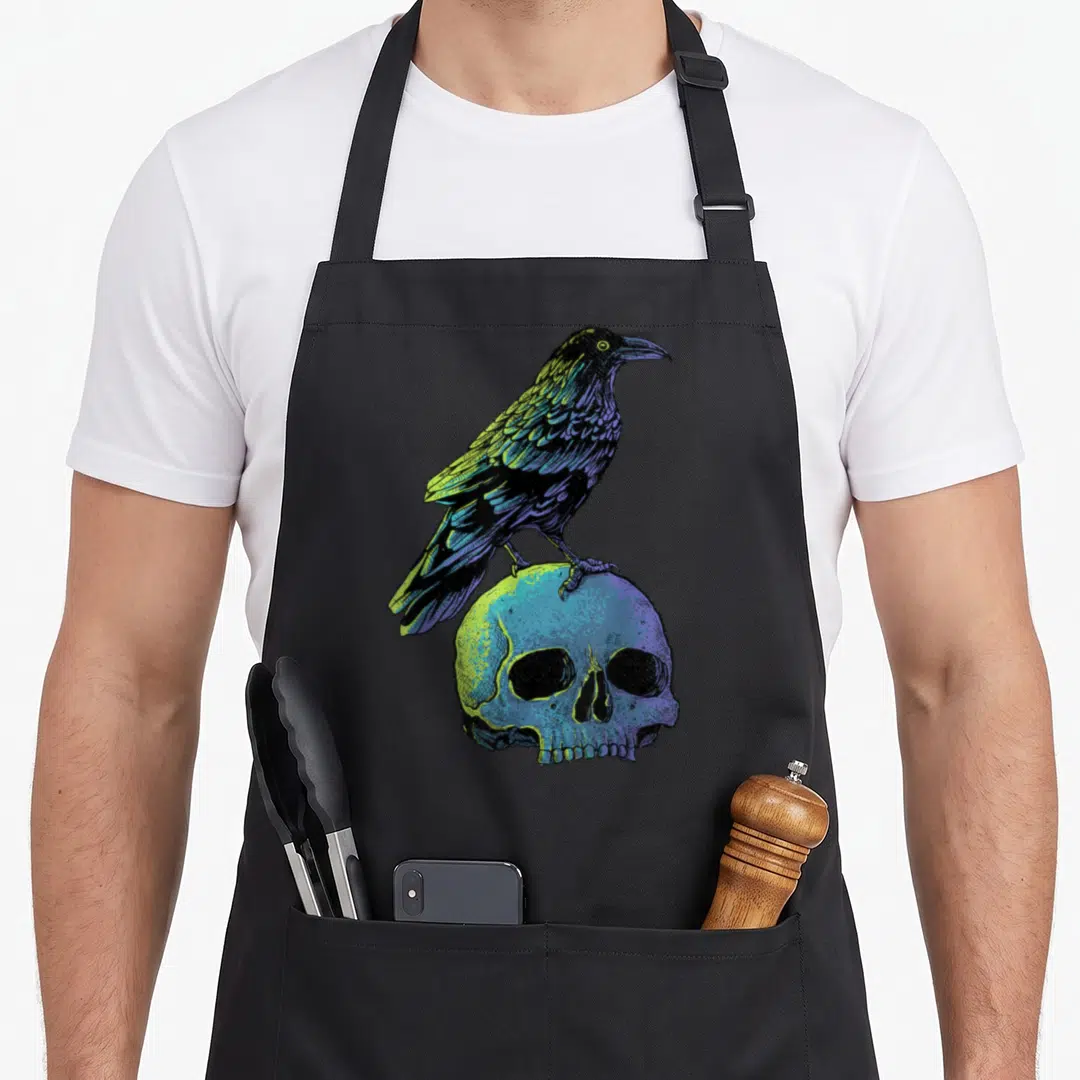 Crow skull halloween apron - Realistic raven in purple and yellow perched on a blue skull — shown on a black cotton canvas apron against a white background