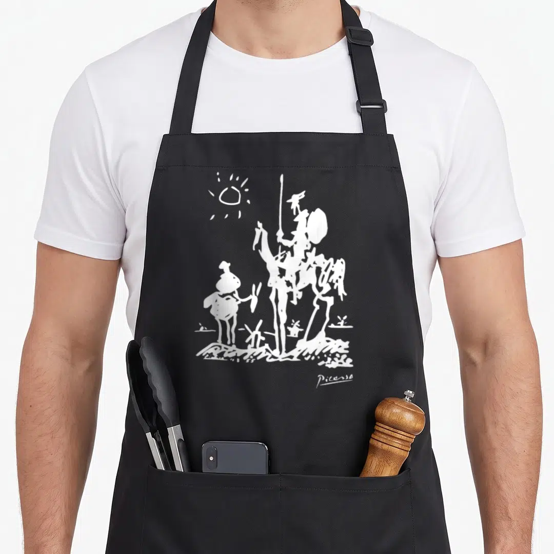 Quixote artwork apron - White line art of two figures, one with a spear, standing under a sun on textured ground — shown on a black cotton canvas apron against a white background