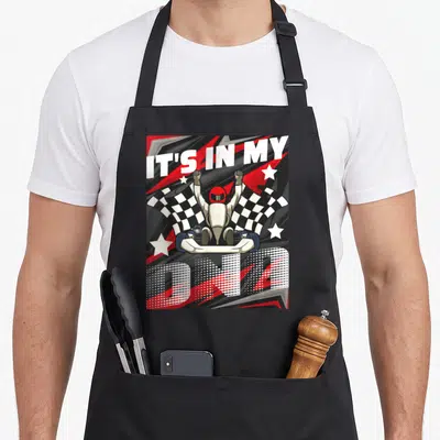 Dna racing apron - Cartoon go-kart driver with raised fists flanked by checkered flags and IT'S IN MY DNA text — shown on a black cotton canvas apron against a white background