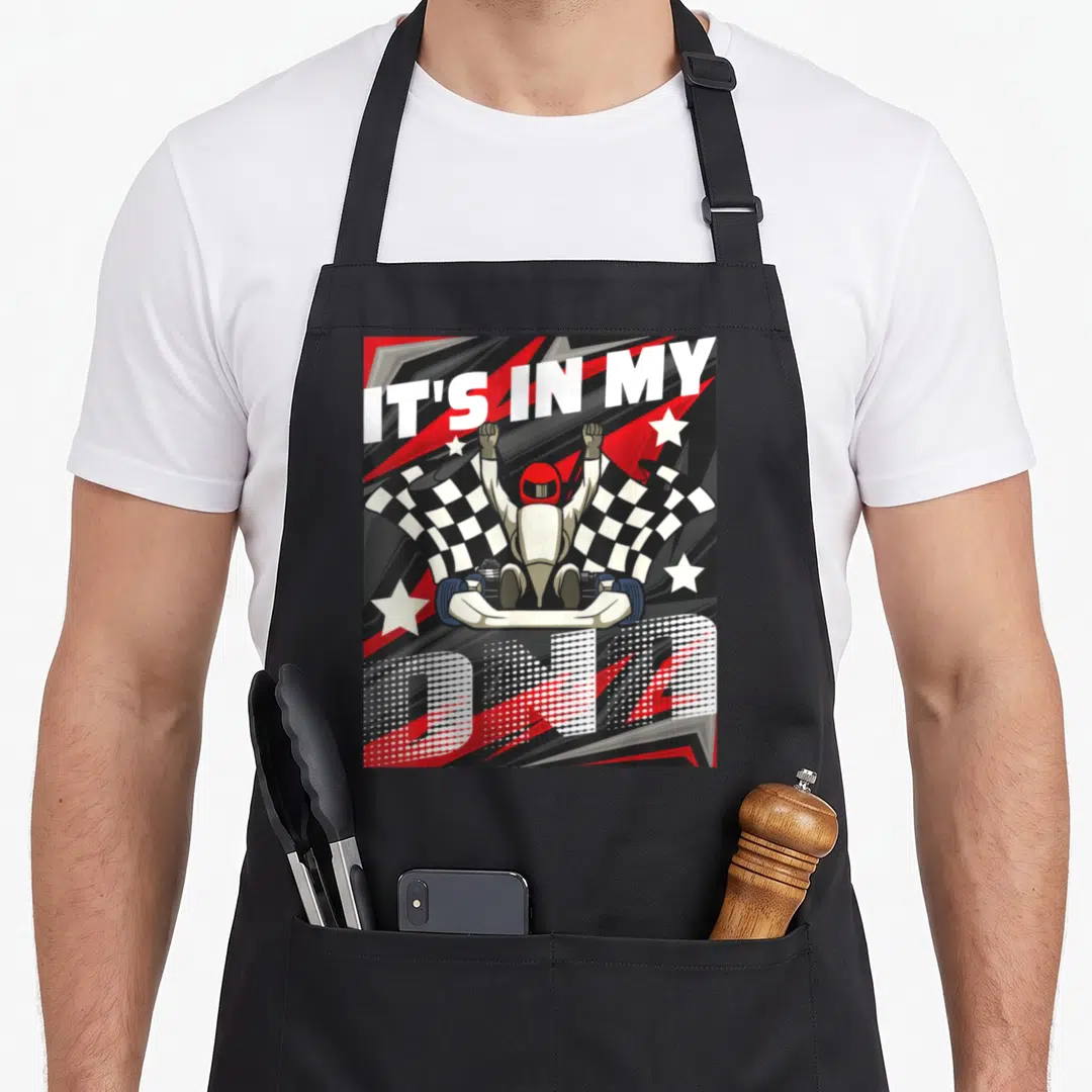 Dna racing apron - Cartoon go-kart driver with raised fists flanked by checkered flags and IT'S IN MY DNA text — shown on a black cotton canvas apron against a white background