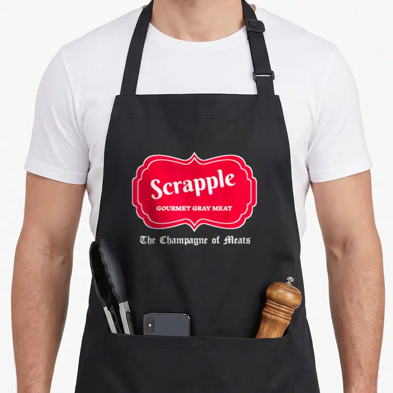 Scrapple champagne apron - Red label reading Scrapple Gourmet Gray Meat with white gothic text below on gray background — shown on a black cotton canvas apron against a white background