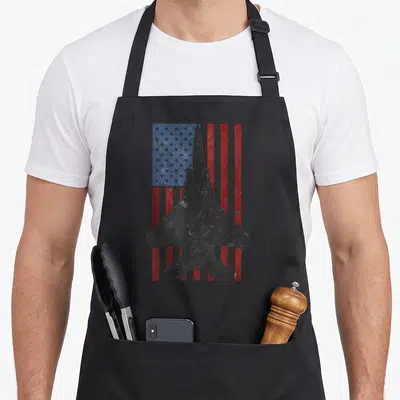 Eagle american flag apron - Black fighter jet silhouette over distressed red white and blue flag pattern — shown on a black cotton canvas apron against a white background