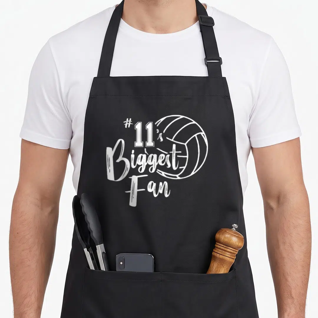 Eleven volleyball dad apron - White #11s Biggest Fan text stacked next to a volleyball outline on canvas — shown on a black cotton canvas apron against a white background