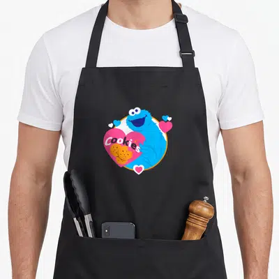 Cookie monster cookies apron - Blue character hugging pink heart with Cookies text and orange biscuits — shown on a black cotton canvas apron against a white background