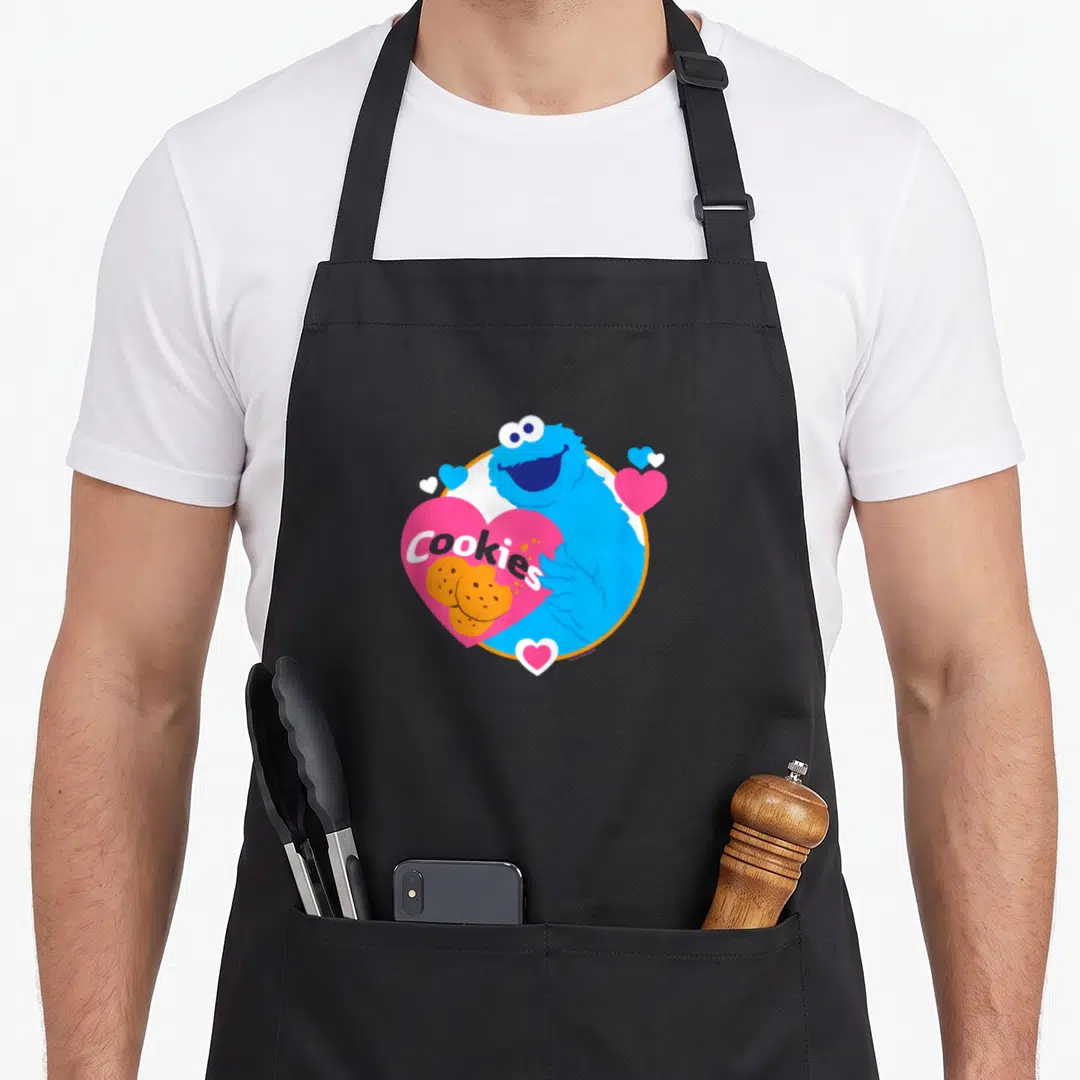 Cookie monster cookies apron - Blue character hugging pink heart with Cookies text and orange biscuits — shown on a black cotton canvas apron against a white background