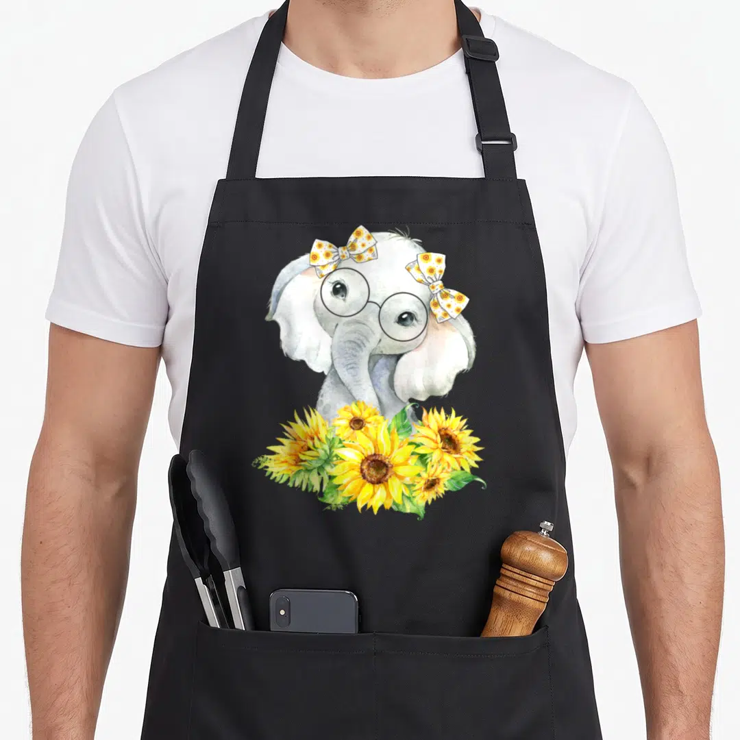 Funny elephant sunflower apron - Grey elephant with glasses and two sunflower bows above yellow flowers in watercolor — shown on a black cotton canvas apron against a white background